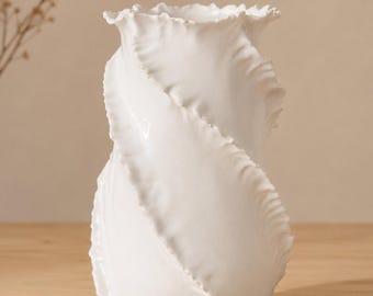 May include: A white ceramic vase with a unique, swirling design. The vase has a ruffled top and a textured, petal-like pattern that wraps around its body. The vase is displayed on a wooden surface, showcasing its elegant form.