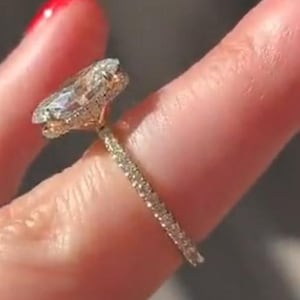 May include: A gold ring featuring a large oval-cut center stone set in a basket setting with small pavé diamonds lining the thin band. The setting includes a hidden halo of small stones beneath the main oval gem, creating a sparkling profile.