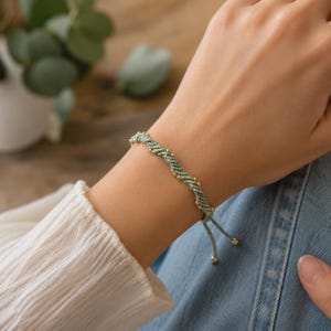 Puede incluir: Una pulsera de cordón verde salvia con un diseño trenzado y retorcido, acentuada con pequeñas cuentas de metal dorado pulido espaciadas uniformemente. La pulsera incluye dos extremos de cordón colgantes terminados con cuentas doradas.