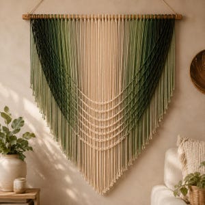 Puede incluir: Un gran tapiz de macramé colgado de un listón de madera, con un diseño en V degradado que transita desde el verde bosque oscuro en los bordes exteriores hasta un color crema claro en el centro, con detalles de cuerdas anudadas.