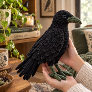 Op de afbeelding: Een gehaakt kraaienfiguur in het zwart met gestructureerde, gelaagde vleugels, een donkergroene snavel en donkergroene poten. De vogel heeft één zichtbaar blauw oog en is gemaakt met complexe garensteken die het uiterlijk van veren nabootsen.