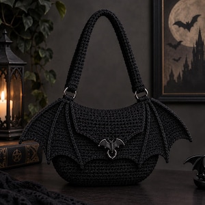 May include: A black crocheted handbag featuring a structured body with prominent bat-wing shaped side panels. The bag includes a single top handle attached with metal rings and a central metal bat-shaped clasp closure on the front flap.