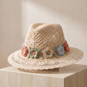 May include: A beige straw fedora hat featuring an intricate open-weave pattern on the crown and brim. A decorative band encircles the base, adorned with crocheted flowers in shades of coral, sage green, and light blue, accented with small beads.