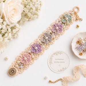 May include: A crocheted bracelet in cream yarn featuring five multi-colored floral motifs in shades of pink, purple, grey, and sage green. The design is accented with white faux pearls, gold beads, and a single brown button closure at one end.