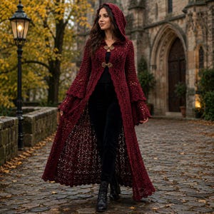 May include: A long, dark red crochet hooded coat featuring intricate lace patterns, wide bell sleeves, and two ornate metallic clasps at the chest. The garment is worn over a black top and black trousers, creating a layered look with a scalloped hem.