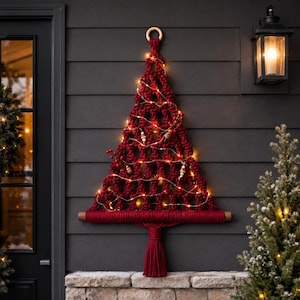 May include: A dark red macrame wall hanging shaped like a Christmas tree, suspended from a wooden ring. The design features intricate knotted cord patterns, a thick horizontal base, a central tassel, and warm white string lights with small beads.
