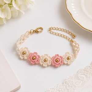 May include: A delicate bracelet featuring five crocheted flower motifs in alternating cream and soft pink colours, each centred with a small gold bead. The band is crafted from cream-coloured lace, finished with a gold-toned lobster clasp and chain.