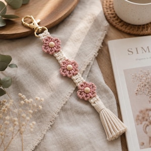 May include: A macrame keychain featuring three dusty pink crocheted flowers with central wooden beads, attached to a gold-toned metal lobster clasp. The off-white cord is knotted in a decorative pattern and finished with a matching fringe tassel.