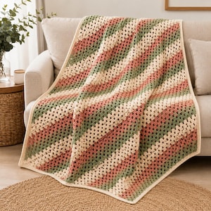 May include: A crochet blanket featuring a diagonal granny square pattern with alternating stripes of cream, sage green, and dusty rose yarn. The rectangular textile has a finished border in cream and displays a consistent open-work stitch texture.