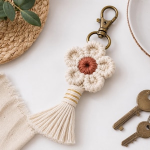 May include: A macrame keychain featuring a flower shape with off-white petals and a rust-colored center. The flower is attached to a bronze-toned metal lobster clasp and includes a long, off-white fringe tassel with three horizontal gold threads.