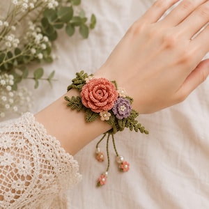 May include: A handcrafted crochet bracelet with a coral rose, purple and beige flowers, and green leaves. The bracelet has dangling pearl and flower accents. A delicate, feminine accessory, perfect for adding a touch of floral charm.
