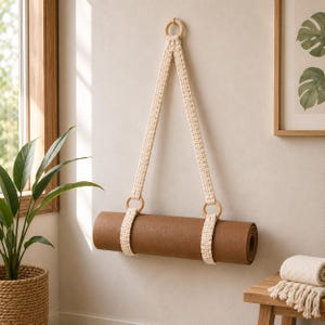Peut inclure: Un tapis de yoga marron suspendu par une sangle en macramé couleur crème avec des motifs noués complexes et trois anneaux en bois lisse. La sangle forme un triangle, avec deux anneaux fixant le tapis et un anneau supérieur pour la suspension.
