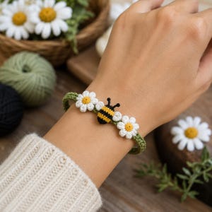 Könnte beinhalten: Ein grünes Häkelarmband mit einem zentralen Hummelmotiv mit gelben und schwarzen Streifen, flankiert von zwei weißen Gänseblümchen mit gelben Mitten. Das Armband ist aus Garn gefertigt und zeigt ein detailliertes, strukturiertes Blumendesign.
