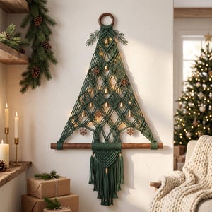May include: A wall-hanging macrame Christmas tree in dark green cord, featuring a wooden ring at the top and a horizontal wooden dowel at the base. The design includes integrated string lights, small gold-toned bells, and wooden snowflake accents.