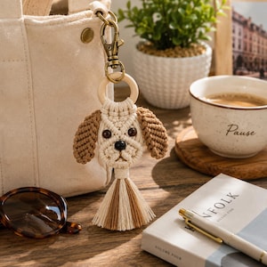 Può includere: Un portachiavi a forma di cane in macramè con viso color crema, orecchie marroni, occhi a perla neri e naso a perla nero. La testa del cane è fissata a un anello in legno chiaro e a un moschettone in metallo bronzo, con una nappa crema e marrone.