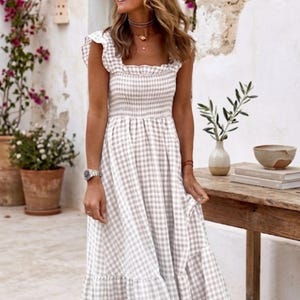 Puede incluir: Un vestido largo hasta los pies con un patrón de cuadros vichy en color beige claro y blanco. El vestido presenta un escote cuadrado, mangas con volantes, un cuerpo fruncido y una falda fluida de varios niveles con dobladillo.