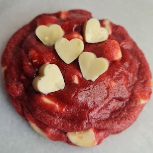Peut inclure: Un cookie de velours rouge avec des morceaux de chocolat blanc en forme de cœur. Le cookie est rond et a une surface légèrement texturée. Les cœurs en chocolat blanc sont dispersés sur le dessus.