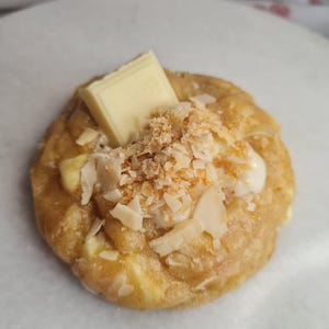 Peut inclure: Une pâtisserie unique, cuite au four, de couleur brun doré, garnie de chocolat blanc, de noix de coco râpée et d'une touche de crème. La pâtisserie repose sur une surface blanche, mettant en valeur les textures et les couleurs du dessert.