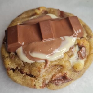 Peut inclure: Gros plan sur un cookie fraîchement cuit, garni d'une barre chocolatée et d'un filet de chocolat blanc. Le cookie est brun doré avec des pépites de chocolat visibles et une texture moelleuse. La barre chocolatée est brun foncé.
