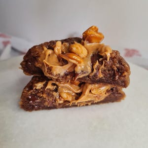 Peut inclure: Un cookie coupé en deux et empilé, garni de beurre de cacahuète et de cacahuètes. Le cookie est brun foncé, avec un aspect doux et texturé. Du beurre de cacahuète est étalé entre les moitiés et garni de cacahuètes entières.