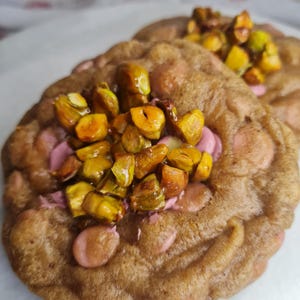 Peut inclure: Deux cookies fraîchement cuits avec un extérieur brun et une garniture rose. Les cookies sont garnis de pistaches hachées. L'image est une vue rapprochée des cookies, mettant en valeur leur texture et leurs ingrédients.