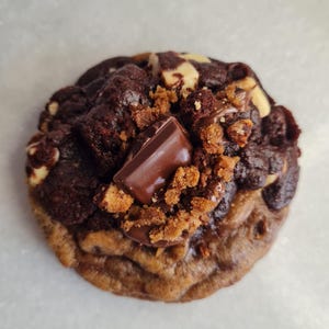 May include: A close-up of a gourmet chocolate cookie. The cookie has a golden-brown base with a dark chocolate topping. It is garnished with chunks of chocolate, nuts, and cookie crumble.