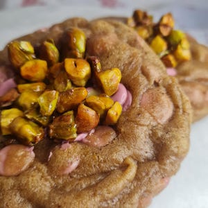 Può includere: Primo piano di due biscotti marroni con glassa rosa e pistacchi tritati. I biscotti hanno una consistenza leggermente irregolare e sono ricoperti da una generosa quantità di noci verdi e marroni. La glassa rosa è visibile al centro.