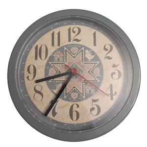 May include: A round wall clock with a gray frame and a beige face featuring black numerals. The center displays a star pattern with hearts, houses, and birds in blue and tan. It has black hour and minute hands and a thin red second hand.
