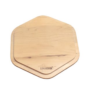 May include: A light-coloured wooden serving board with a hexagonal shape and rounded corners. The surface displays a natural wood grain pattern with visible knots. The brand name LONGABERGER WOODCRAFTS is stamped in black ink on the lower section.