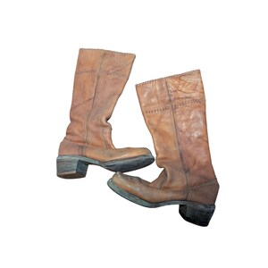 Puede incluir: Un par de botas de cuero a media pantorrilla en color marrón claro. Estas botas presentan una punta cuadrada, un tacón de bloque apilado y detalles de costuras decorativas a lo largo de la caña. El cuero tiene una textura desgastada con costuras visibles y suelas oscuras.