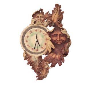 May include: A wall clock featuring a circular face with black hands and numbers 3, 6, 9, and 12. The clock is surrounded by sculpted brown and green foliage and two faces with closed eyes, integrated into the leaf-like frame design.