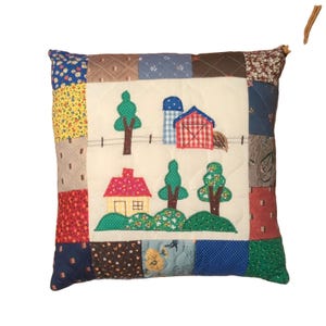 Peut inclure: Un coussin carré en patchwork avec un panneau central crème orné d'applications en tissu représentant une grange rouge, un silo bleu, une maison jaune et des arbres verts. La bordure est composée de divers carrés de tissu à motifs colorés.