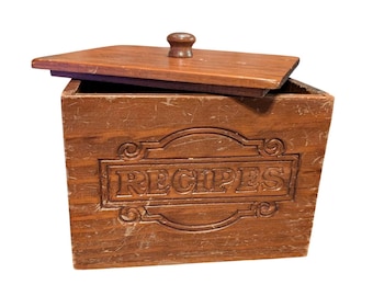 May include: A rectangular brown wooden recipe box with a matching lid featuring a small round knob. The front of the box has the word RECIPES embossed in a decorative frame. The wood shows visible grain and signs of wear on the surface.