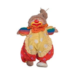 Puede incluir: Muñeca de payaso vintage con cara pintada, pelo de hilo marrón y cuello naranja con volantes. La muñeca viste un mono amarillo con lunares blancos y pantalones rojos con estampados florales. Las mangas son de color arcoíris.