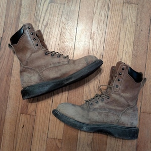 Puede incluir: Un par de botas de trabajo de cuero marrón con diseño de caña alta, cuellos acolchados negros, ojales de metal oscuro y suelas gruesas de goma negra. Las botas presentan un acabado texturizado desgastado y cordones marrones a juego.