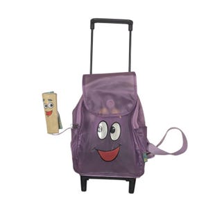 May include: A purple rolling backpack featuring a cartoon face with large white eyes and a smiling mouth on the front flap. The bag has a retractable black handle, wheels at the base, and a small attached pouch shaped like a smiling map character.