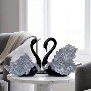Puede incluir: Dos figuras de cisnes con cuellos y cabezas de color negro liso y alas plateadas metálicas texturizadas. Los cisnes están colocados uno frente al otro, mostrando patrones de plumas detallados en sus alas y cuellos curvos sobre una mesa blanca.