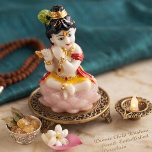 May include: A porcelain figurine of a child playing a flute, seated on a pink lotus base atop a round bronze-toned stand. The figure has black hair with a peacock feather, red and yellow clothing, and gold jewellery. Text reads: Divine Child Krishna Hand-Embellished Porcelain.