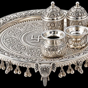 May include: A silver-coloured metal puja thali set featuring an ornate circular tray with elephant-shaped feet and hanging bells. The tray displays intricate floral engravings and a central swastika symbol, accompanied by two lidded containers, a small pot, and a diya lamp.