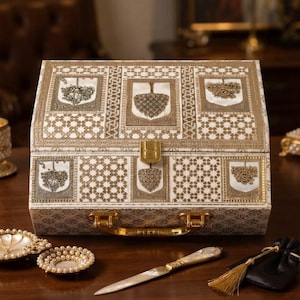 May include: A rectangular white and gold storage box with a hinged lid and a gold metal latch. The exterior features intricate geometric patterns and decorative gold-toned floral motifs. A matching letter opener with a pearlescent handle is included.