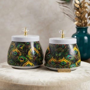 May include: Two rounded storage jars featuring a vibrant peacock feather pattern in shades of green, blue, and gold. Each jar has a white lid topped with a clear crystal-like knob. A small gold-toned rectangular tag labelled Artisan Peacock is attached.