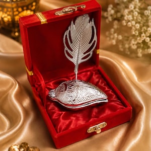 Puede incluir: Un objeto de metal plateado con forma de concha y una pluma vertical, presentado dentro de una caja cuadrada forrada de terciopelo rojo con un cierre de metal dorado. El objeto presenta intrincados patrones en relieve en su superficie.