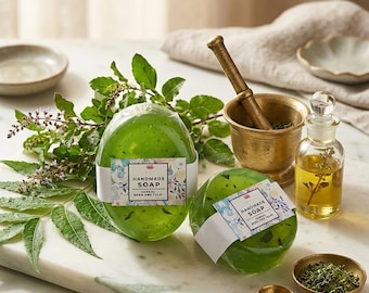 Könnte beinhalten: Zwei durchscheinende grüne ovale Seifenstücke, eingewickelt in weiße Papierbanderolen mit Blumenmuster und dem Text "HANDMADE SOAP HERBAL NEEM AND TULSI". Die Seife ist mit grünen Blättern, einem Messingmörser mit Stößel und Öl arrangiert.