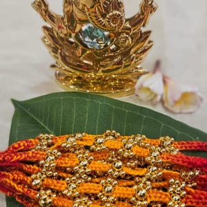 May include: A braided bracelet featuring vibrant orange and red threads interwoven with numerous small, reflective gold-colored metallic beads. The bracelet is positioned on a large green leaf, with a gold-toned Ganesha figurine in the background.