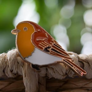 May include: A metal enamel pin shaped like a bird, featuring a bright yellow head, a reddish-brown wing and back, and a white chest. The bird design is outlined in a metallic gold tone, showcasing detailed wing feathers and a small pointed beak.