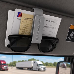 May include: A gray car visor organizer with the text "PinaPrecise 3D" holding documents and black sunglasses. A warning sticker is visible. The background shows trucks on a highway.