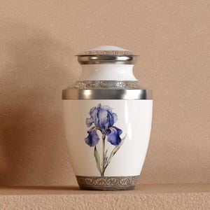May include: A white cremation urn featuring a blue iris flower illustration on the front. The urn has a silver-toned lid and base, both decorated with an intricate engraved scrollwork pattern. The body of the urn is smooth and glossy white.