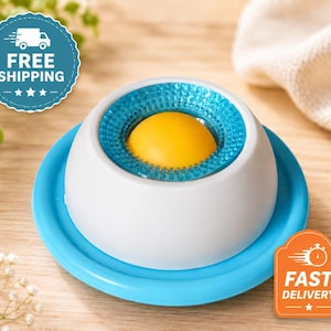 Puede incluir: Un objeto en forma de c&uacute;pula blanca con una esfera central de color amarillo brillante rodeada por un anillo azul transl&uacute;cido y texturizado. El objeto se asienta sobre una base circular plana de color azul. Acabado exterior blanco mate.