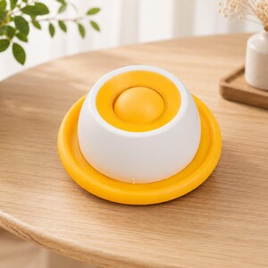 Puede incluir: Un objeto redondo en forma de c&uacute;pula con una base de color amarillo brillante y una secci&oacute;n superior blanca. El centro de la c&uacute;pula blanca contiene un elemento circular amarillo similar a un bot&oacute;n. El dise&ntilde;o es liso y sim&eacute;trico.