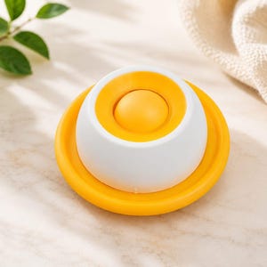 Puede incluir: Un objeto circular en forma de c&uacute;pula que presenta una base exterior blanca con un borde amarillo brillante y un bot&oacute;n central circular amarillo. El dise&ntilde;o es sim&eacute;trico, con un acabado liso y mate, y descansa sobre una superficie plana clara.